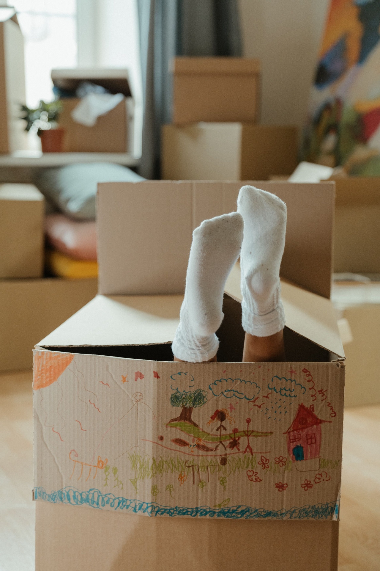 Child's feet sticking out of a crayon-decorated moving box, surrounded by packed cardboard boxes in a sunlit home